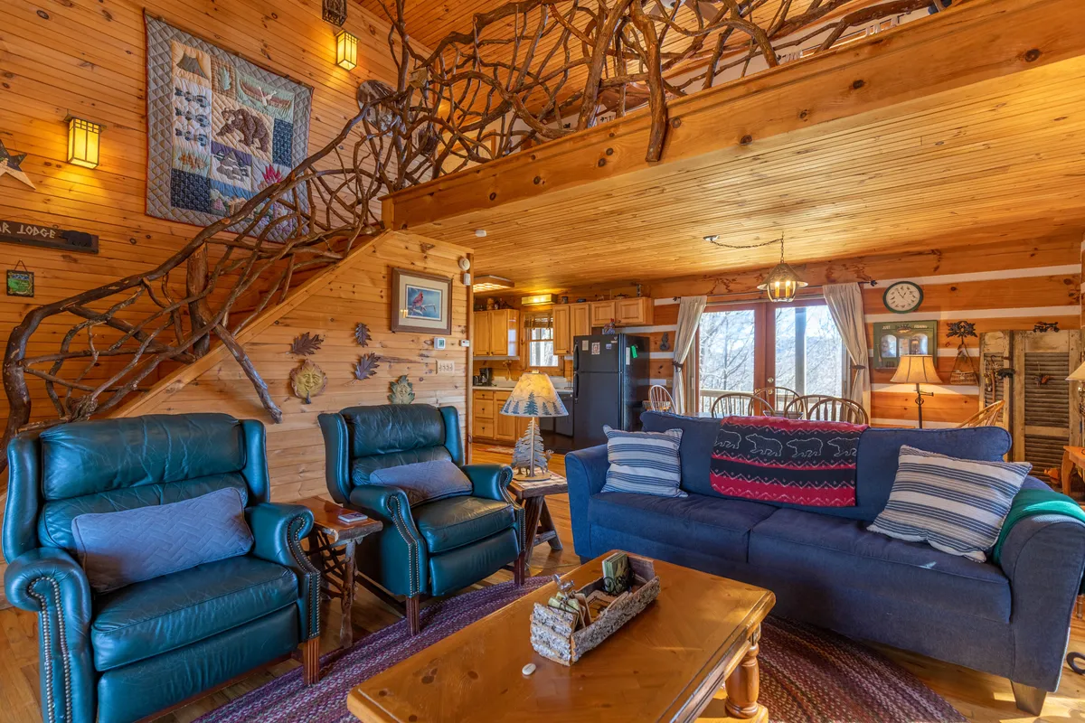 This image features a warm and welcoming living space with a wooden staircase, a blue sofa, leather armchairs, a coffee table with artful decor, and inviting wall art. The space is bathed in natural light, offering a glimpse of mountain charm and cozy relaxation.
