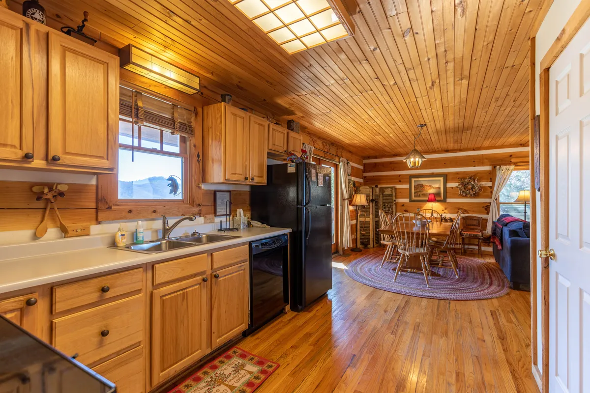 A cozy view looking across the kitchen sink and countertops toward a dining table set for group gatherings, with the living area and a plush couch visible in the background. Warm lighting and wood tones create an inviting atmosphere for family meals and conversation.