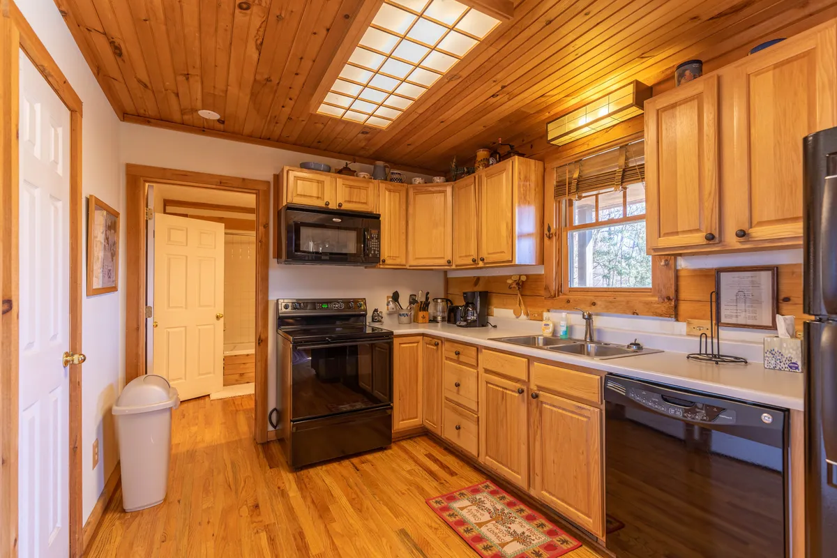 This image features a kitchen with wooden cabinetry and black appliances, bathed in natural light from a nearby window. The scene is warm and inviting, with a rug on the floor and hints of activity such as cooking and gathering.