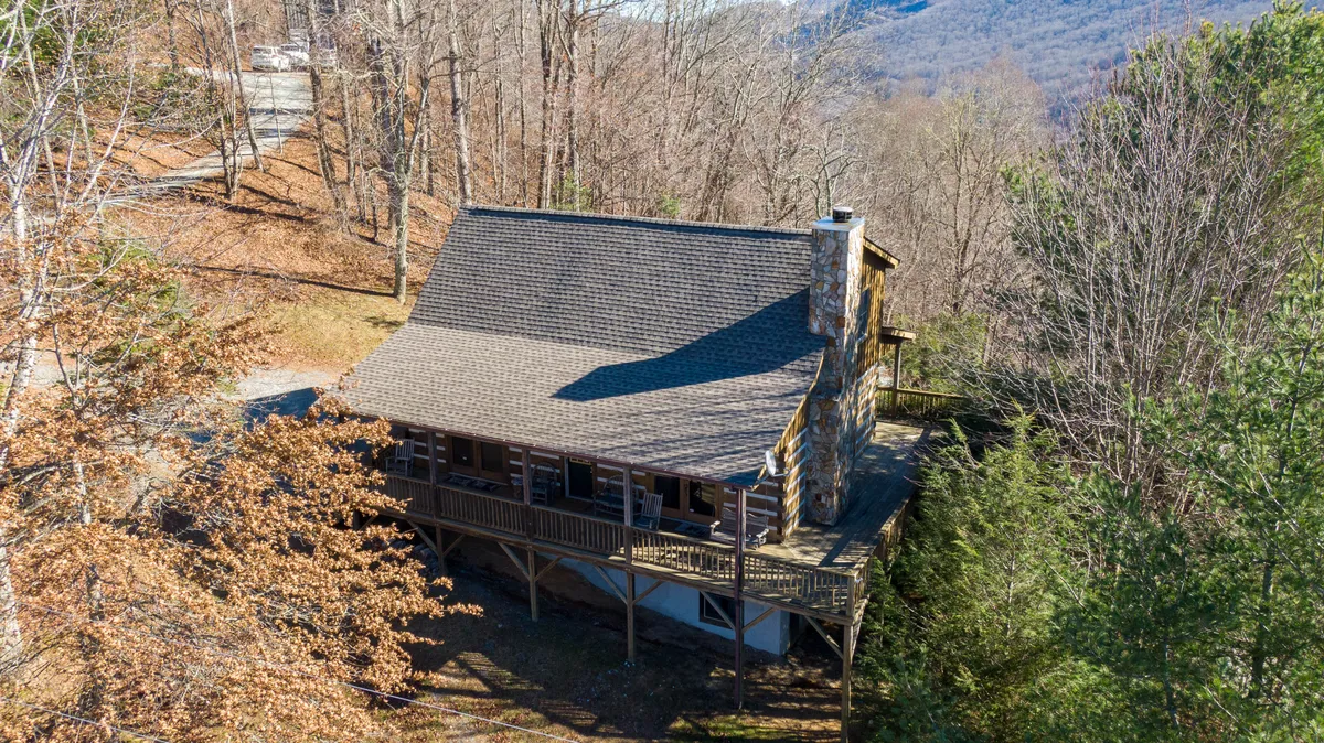 This image captures the expansive wooden wrap-around deck of the log cabin, framed by a stately stone chimney and surrounded by lush trees. The deck offers seating and panoramic views, inviting guests to relax outdoors while enjoying the tranquil mountain atmosphere.