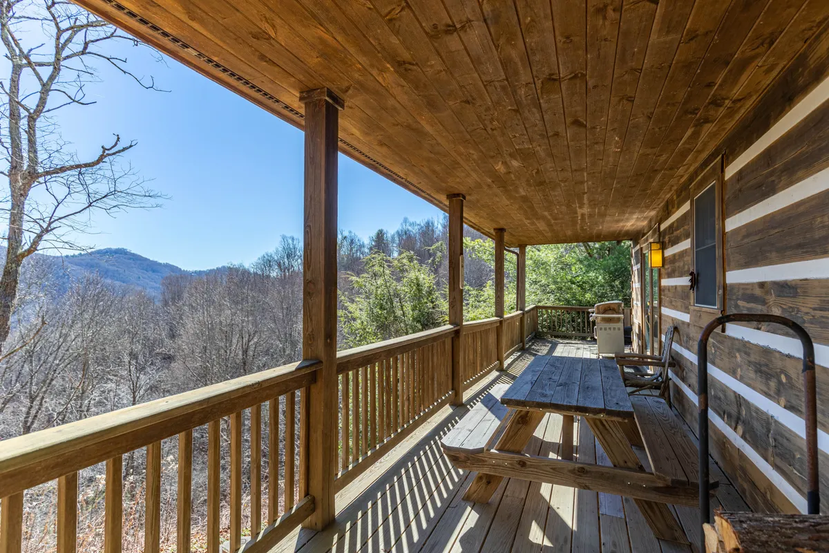The deck's wooden picnic table is positioned to take full advantage of breathtaking mountain views. The sturdy railing and log cabin structure frame the scene, suggesting an inviting spot for family meals or morning coffee.
