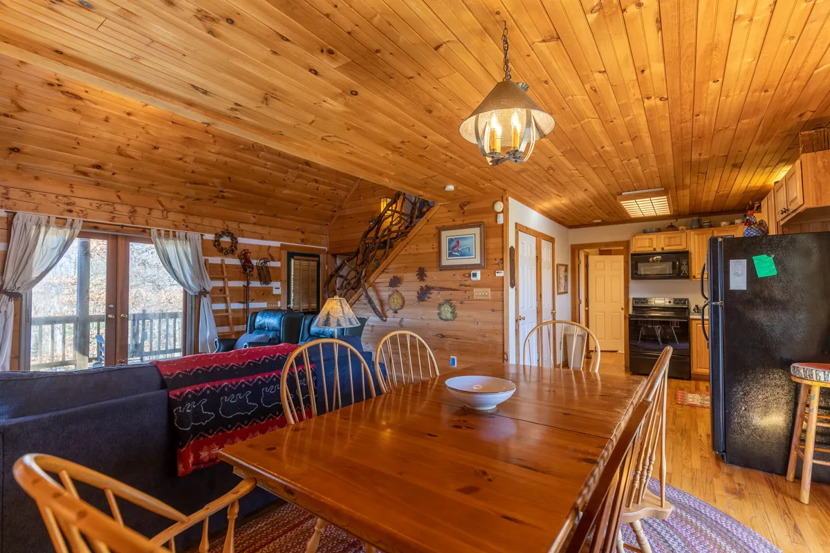 A wide-angle shot of the unified dining and kitchen area, showcasing a wooden dining table, log cabin walls, large windows with scenic views, a staircase with a distinctive twig-style banister, and a well-equipped kitchen with modern appliances.