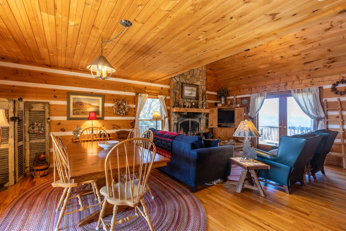 This image captures a warm, inviting open-plan living and dining area featuring a wooden dining table for six, a stone fireplace, large windows offering splendid mountain views, and a cozy seating area. The room is filled with natural light, highlighting the harmonious blend of comfort and rustic elegance.