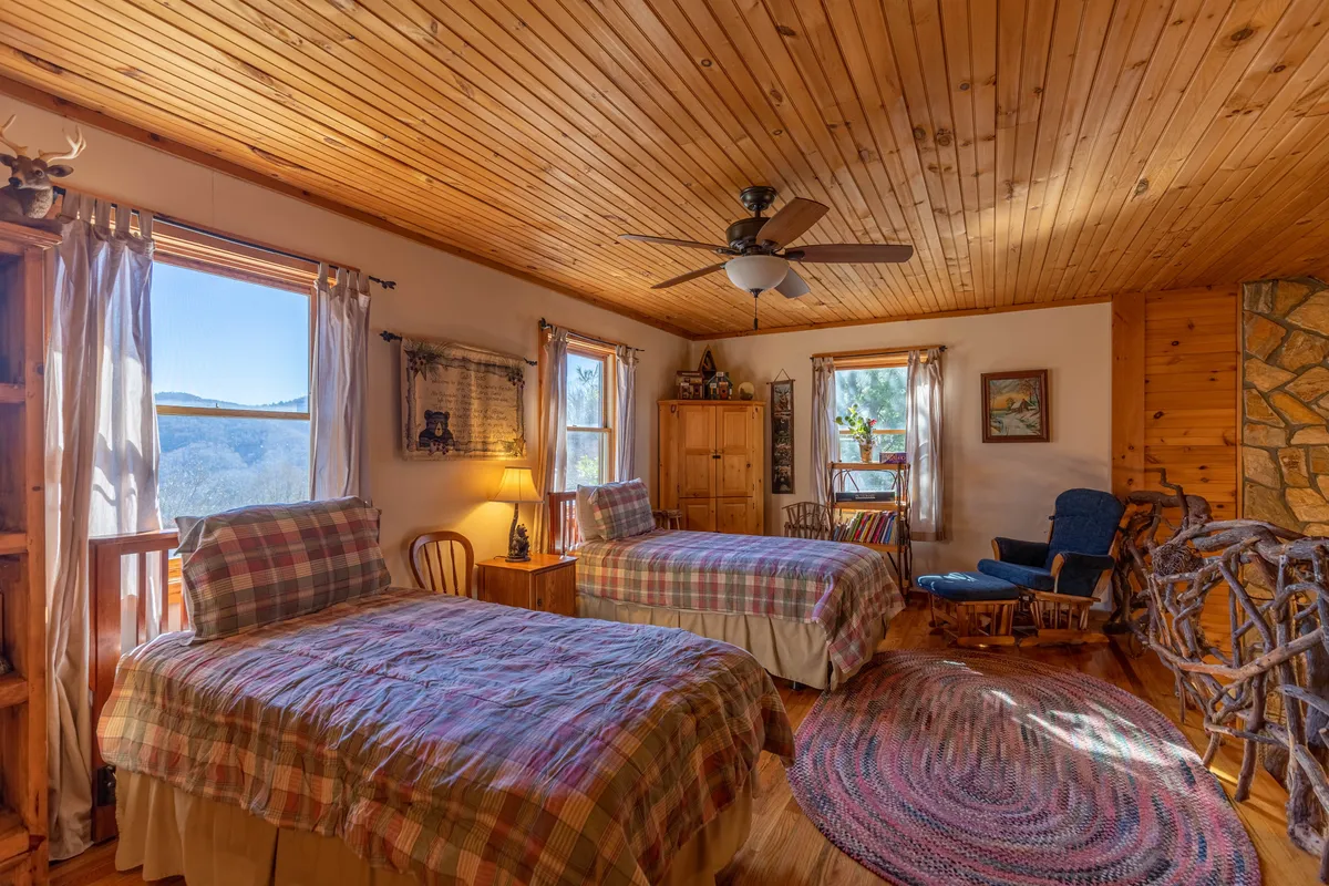 A cozy loft space with two twin beds adorned in plaid bedding, a rustic wooden ceiling and walls, large windows with inviting mountain views, and charming rustic furniture.