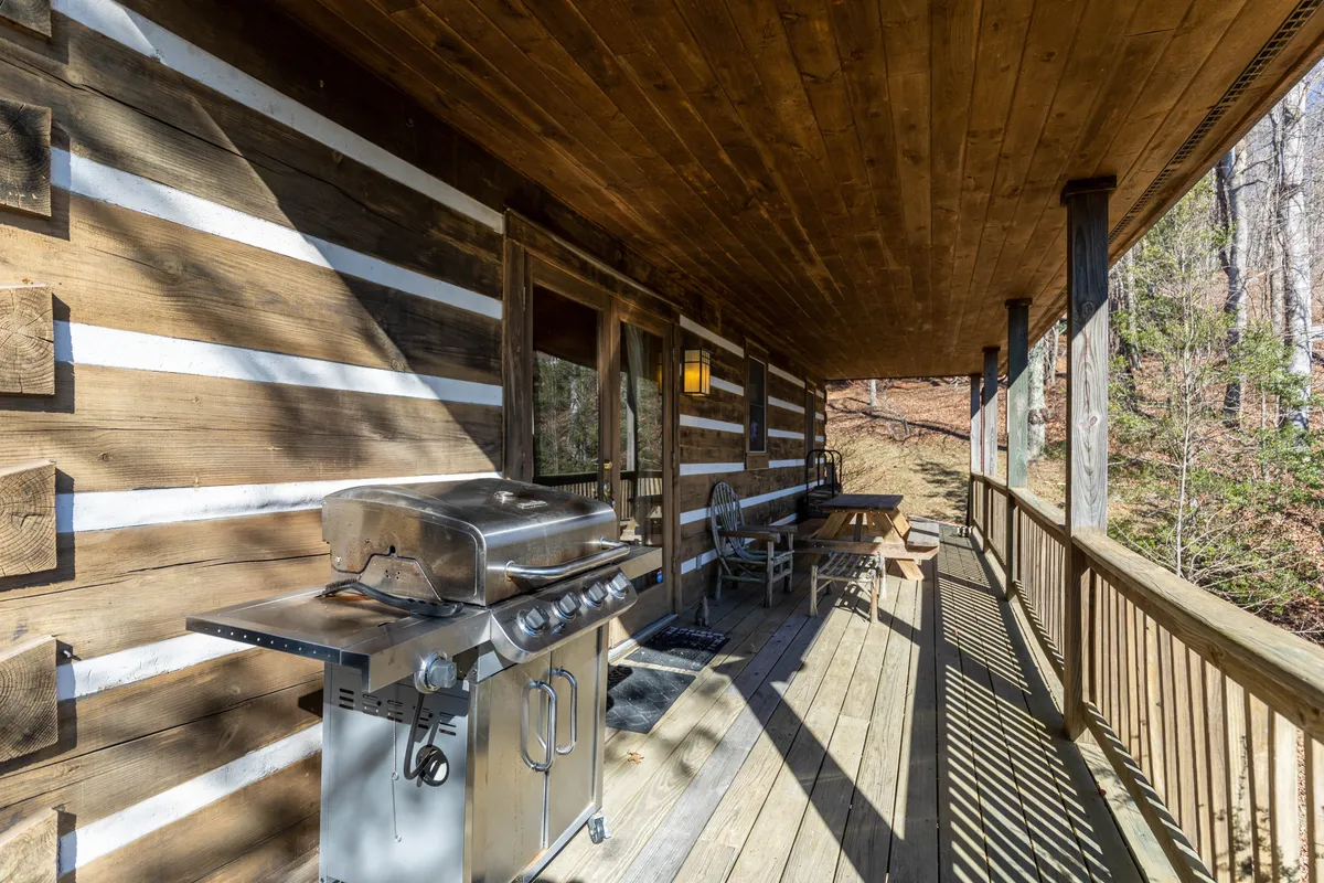 A rustic wooden deck scene featuring a prominent gas grill, outdoor seating, and the characteristic log walls of the cabin. The space is set up for barbecuing, socializing, and relaxing, all with views of the surrounding natural landscape.