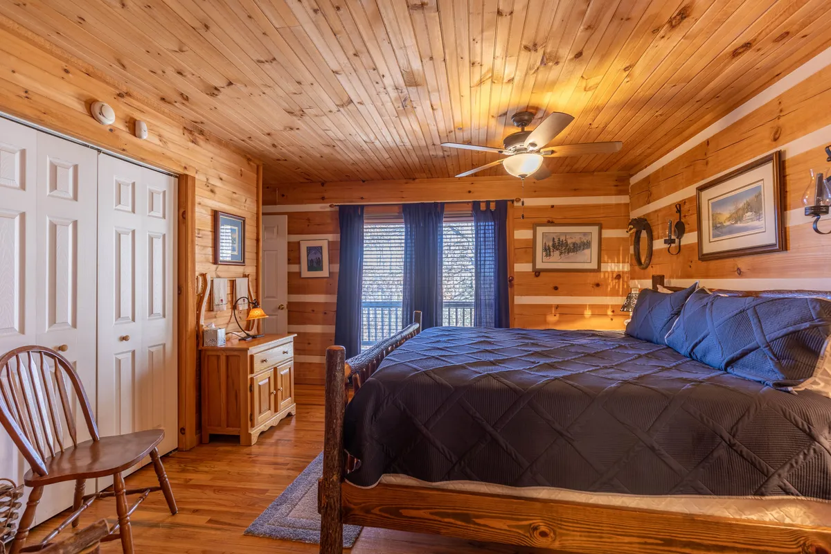 This image showcases a warmly lit bedroom with dark bedding, wooden walls, large windows draped with curtains, and wooden furniture. The tranquil setting exemplifies the peace and comfort expected in a main-level King bedroom.