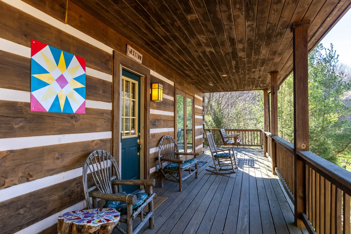 The hero image shows the inviting exterior of the Windsong cabin perched on a mountain top, featuring a welcoming blue door, a vibrant quilt square adorning the wall, and classic wooden rocking chairs on the porch. The setting exudes warmth and comfort, with plenty of space to relax, take in mountain sunsets, and enjoy moments with friends or family.