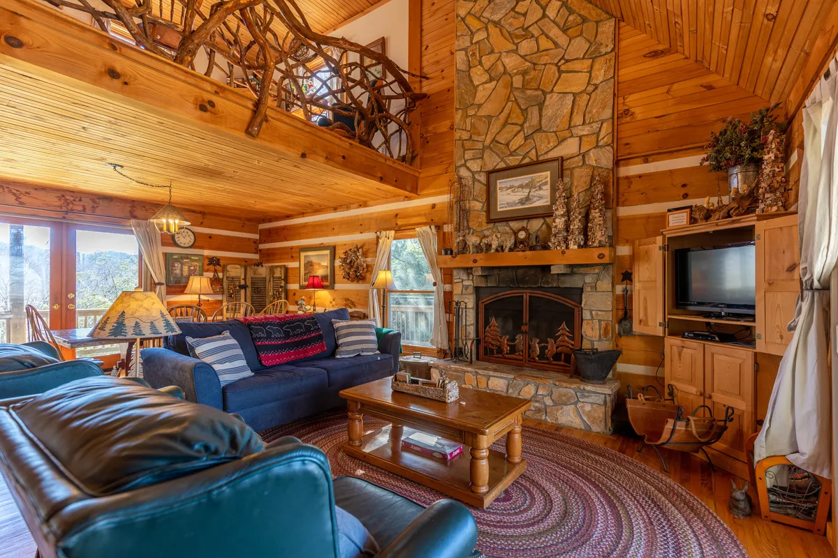 A rustic living room with a prominent stone fireplace, wooden beams, comfy seating, and large windows allowing nature to spill inside. The decor accentuates warmth and mountain charm.